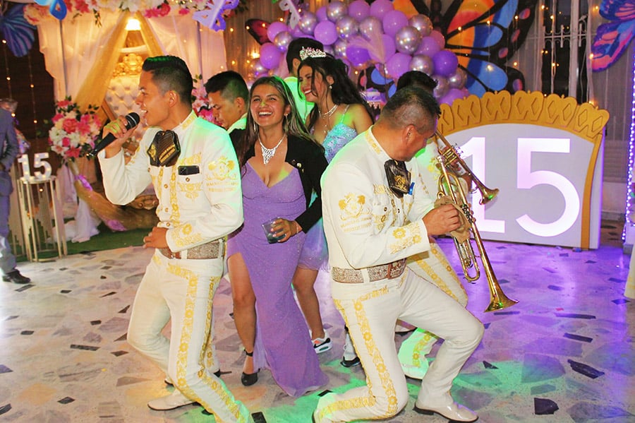 Mariachis en trajes blancos con bordados dorados animando una fiesta de quinceañera, cantando y tocando trompeta mientras interactúan con invitadas en vestidos violeta frente a una decoración festiva con globos y números "15" iluminados.