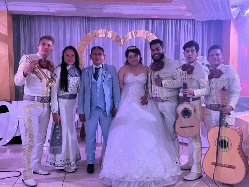 Mariachi Juvenil Fernández posando junto a los recién casados en una boda, músicos con trajes blancos tradicionales sosteniendo sus instrumentos mientras los novios lucen elegantes en el centro, con decoración en tonos claros y románticos.